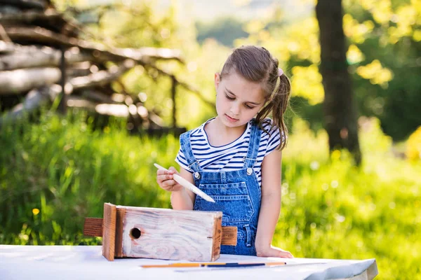 Una bambina con un pennello all'esterno, dipingendo una casetta di legno per uccelli . — Foto Stock