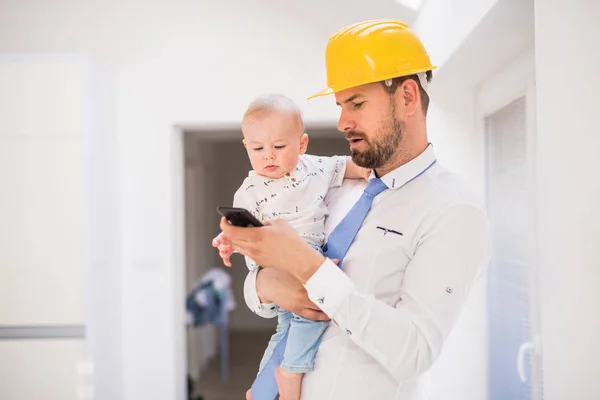 Padre con camicia, cravatta, casco e smartphone che tiene in braccio un bambino a casa . — Foto Stock