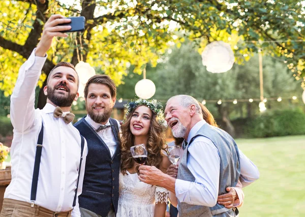 Mariée, marié et invités avec smartphone prenant selfie à l'extérieur à la réception de mariage . — Photo