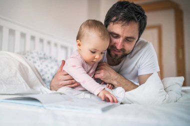 Babası yürümeye başlayan çocuk kızla yatmadan evde yatakta kitap okuma.
