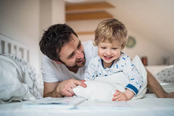Far med småbarn pojke läser en bok på säng hemma vid sänggåendet. — Stockfoto