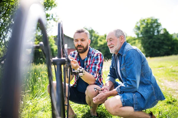 Ein erwachsener Hipster-Sohn und ein älterer Vater reparieren an einem sonnigen Tag draußen ein Fahrrad. — Stockfoto