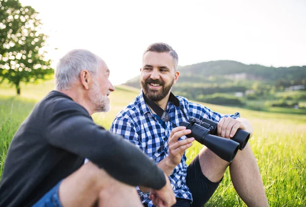 Un hijo hipster adulto con prismáticos y padre mayor sentado en la hierba en la naturaleza soleada . — Foto de Stock