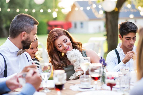 Huéspedes con un pequeño perro sentado en la mesa afuera en el patio trasero . — Foto de Stock