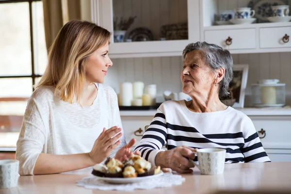 Une grand-mère âgée avec une petite-fille adulte assise à la table à la maison, mangeant des gâteaux . — Photo
