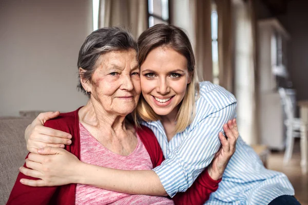 En äldre mormor med en vuxen barnbarn hemma, kramas. — Stockfoto