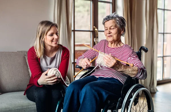 Una abuela anciana en silla de ruedas con una nieta adulta tejiendo en casa . — Foto de Stock