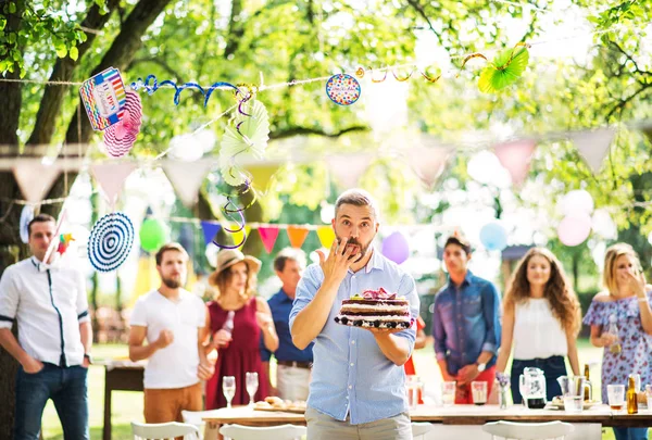 Homem com um bolo em uma celebração familiar ou uma festa no jardim lá fora, lambendo o dedo . — Fotografia de Stock