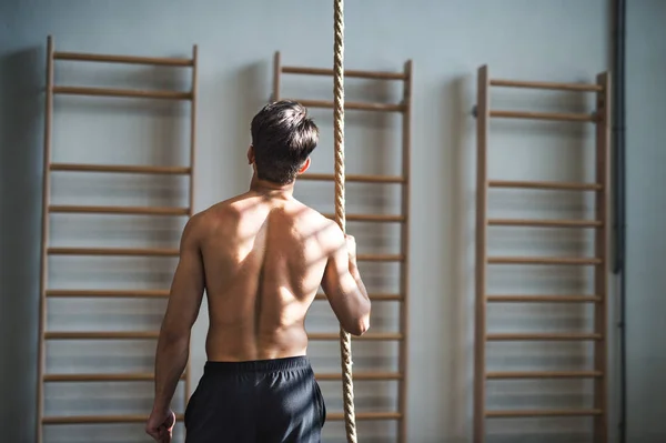 Fit young man in gym standing topless , holding a climbing rope. Rear view. — Stock Photo, Image