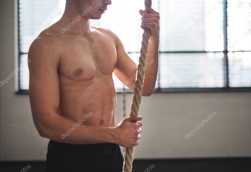 Fit young man in gym standing topless , holding climbing rope.