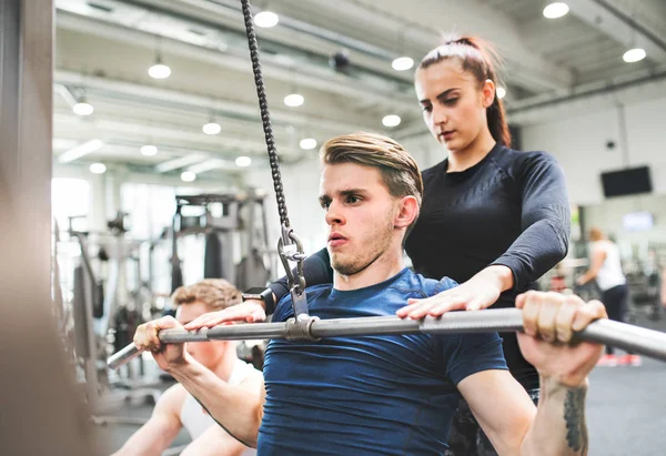 Ung i gymmet träna på pull-down maskin, en personlig tränare bakom honom. — Stockfoto