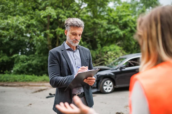 An insurance agent talking to a woman outside on the road after a car accident.