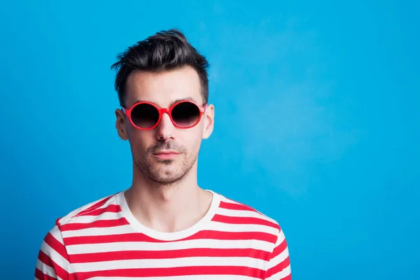 Egy fiatal férfi, napszemüveg, kék háttérrel stúdióban portré. — Stock Fotó