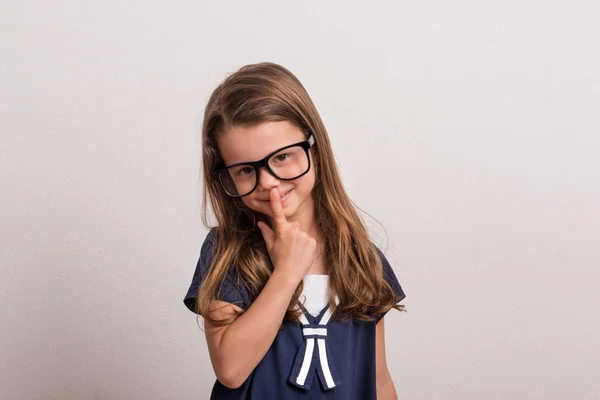 Retrato de una niña pequeña con gafas grandes y un dedo en la nariz en un estudio . —  Fotos de Stock