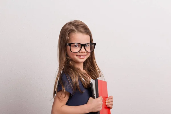 Porträtt av en liten skolflicka med stora glasögon i en studio, hålla anteckningsböcker. — Stockfoto