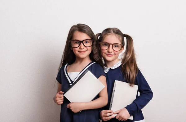 Twee kleine schoolmeisjes met glazen en uniform permanent in een studio, blocnotes houden — Stockfoto