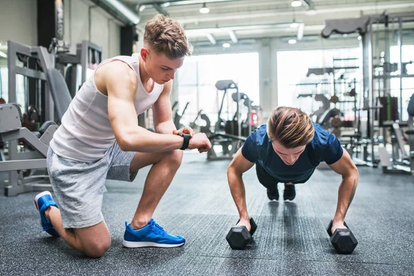 Молодые мужчины в спортзале отжимаются на гантели, измеряют время на умных часах . — стоковое фото