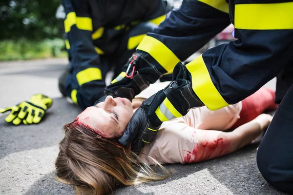 Pompierii de nerecunoscut ajuta o femeie tanara ranita intinsa pe drum dupa un accident . — Fotografie, imagine de stoc