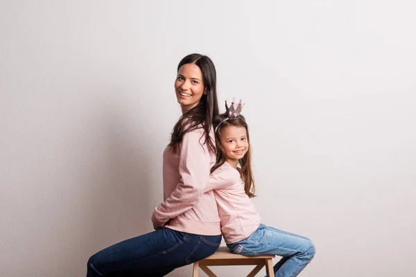En liten flicka med crown pannband och mamman sitter rygg mot rygg i en studio. — Stockfoto