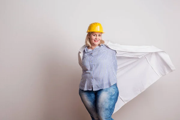 Portrait of an attractive overweight woman with yellow helmet and lab coat in a studio. — Stock Photo, Image