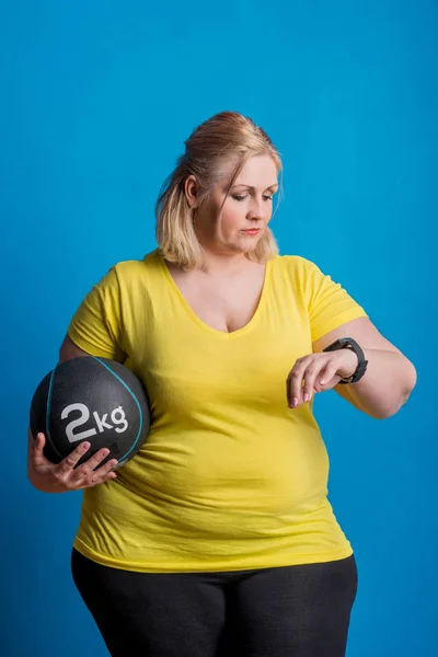 Retrato de una mujer feliz con sobrepeso con smartwatch y bola pesada en el estudio . —  Fotos de Stock