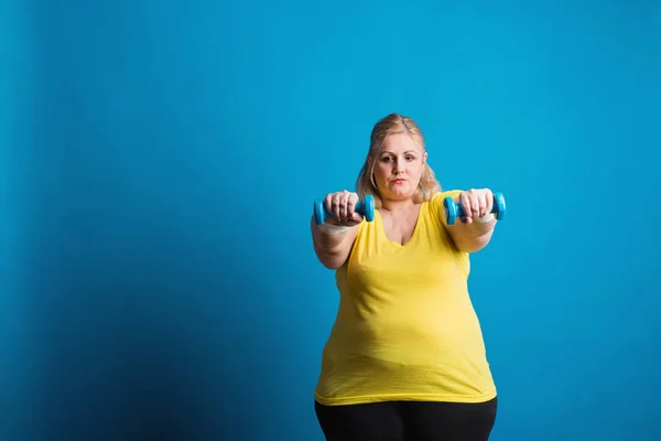 Retrato de una mujer infeliz con sobrepeso y mancuernas en el estudio sobre un fondo azul . —  Fotos de Stock