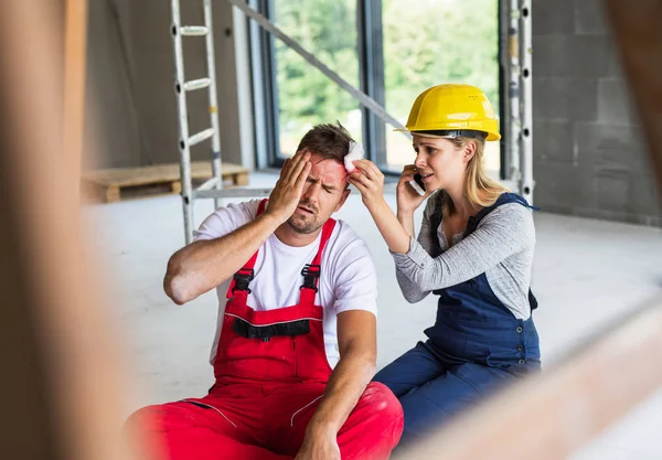 Uma mulher com smartphone ajudando o homem trabalhador após um acidente no local de construção . — Fotografia de Stock