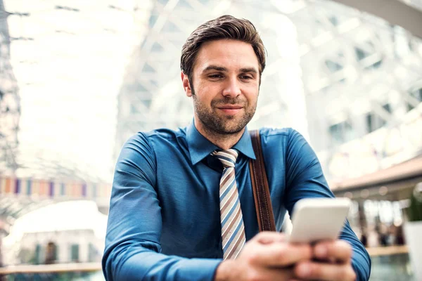 Een jonge zakenman met smartphone staan in een modern gebouw, texting. — Stockfoto