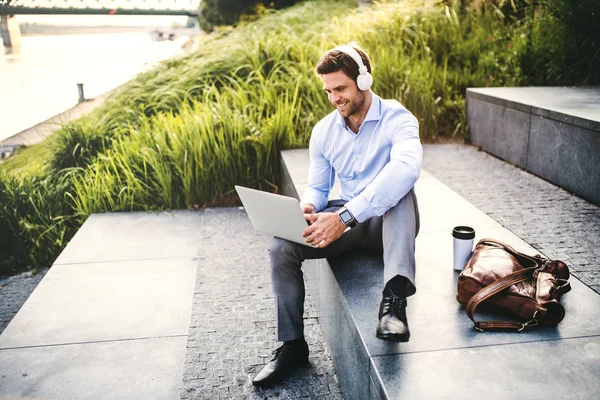 En affärsman med hörlurar som sitter utomhus på trappor, använder laptop. — Stockfoto