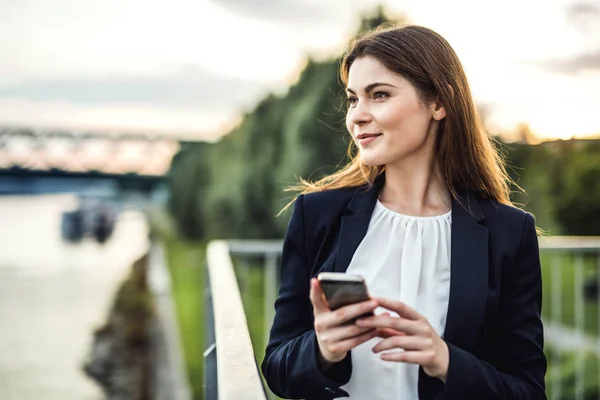 A young businesswoman standing on the river bank, using smartphone. — Stock Photo, Image