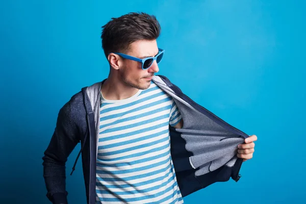 Retrato de um jovem homem feliz com óculos de sol em um estúdio em um fundo azul . — Fotografia de Stock