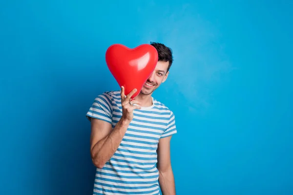 Портрет молодого чоловіка в студії на синьому фоні, що ховається за червоним серцем . — стокове фото
