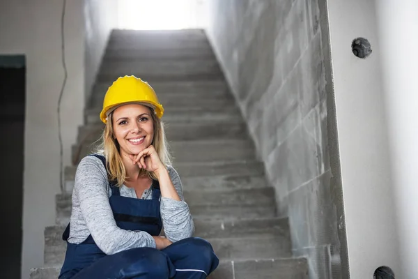 Jonge vrouw werknemer met een gele helm zittend op de trappen op de bouwplaats. — Stockfoto