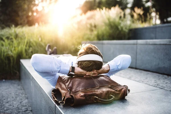 Seorang pengusaha dengan headphone, berbaring di tangga di luar rumah saat matahari terbenam . — Stok Foto