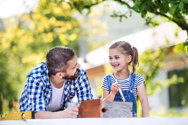 Father with a small daughter outside, painting wooden birdhouse. — Stock Photo, Image