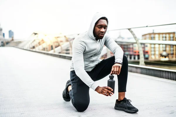 Joven corredor de hombre negro deportivo con botella de agua en el puente en una ciudad, descansando . —  Fotos de Stock