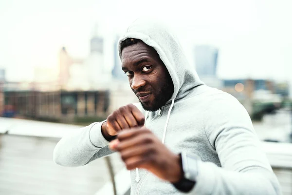 Fiatal active fekete sportoló boksz helyzetben egy városban, kapucnis visel. — Stock Fotó