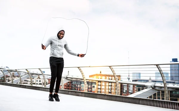 Giovane sportivo nero attivo che salta con una corda in città, indossando una felpa con cappuccio . — Foto Stock