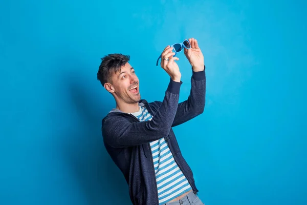 Retrato de um jovem homem feliz com óculos de sol em um estúdio em um fundo azul . — Fotografia de Stock