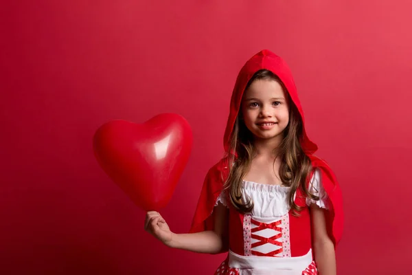 Uma menina pequena em Little Red Riding Hood traje em estúdio em um fundo vermelho . — Fotografia de Stock
