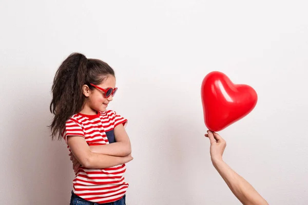 A small girl with red sunglasses and striped T-shirt looking at red heart balloon. — Stock Photo, Image