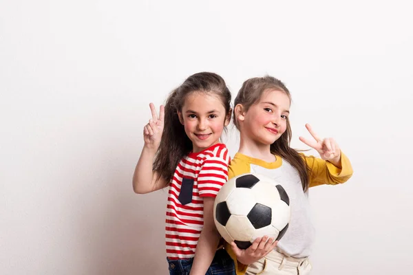 Meninas pequenas com uma bola de futebol em pé em um estúdio, mostrando sinal de vitória . — Fotografia de Stock