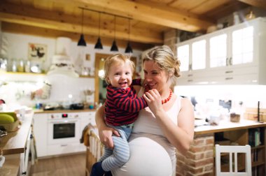 Beautiful pregnant woman carrying a toddler boy in the kitchen at home. clipart
