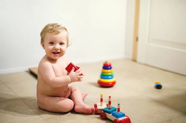 Un niño desnudo sentado en el suelo en casa, jugando con juguetes de madera . — Foto de Stock