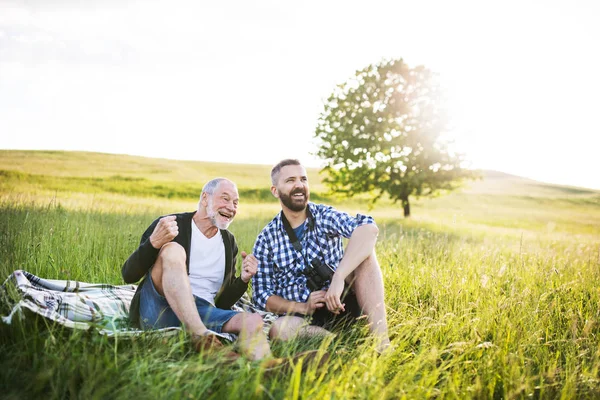 Un hijo hipster adulto con padre mayor sentado en la hierba al atardecer en la naturaleza . —  Fotos de Stock