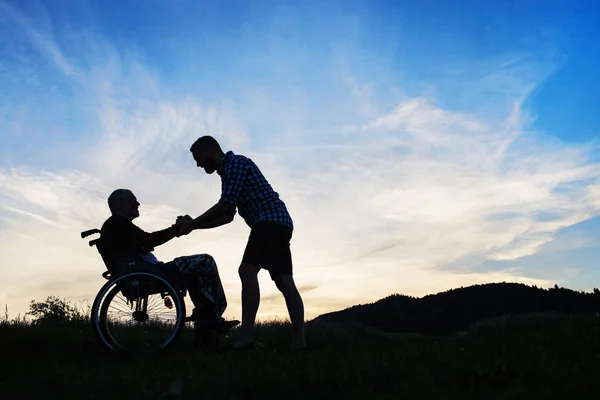 Une silhouette de fils adulte regardant son père en fauteuil roulant dans la nature au coucher du soleil . — Photo