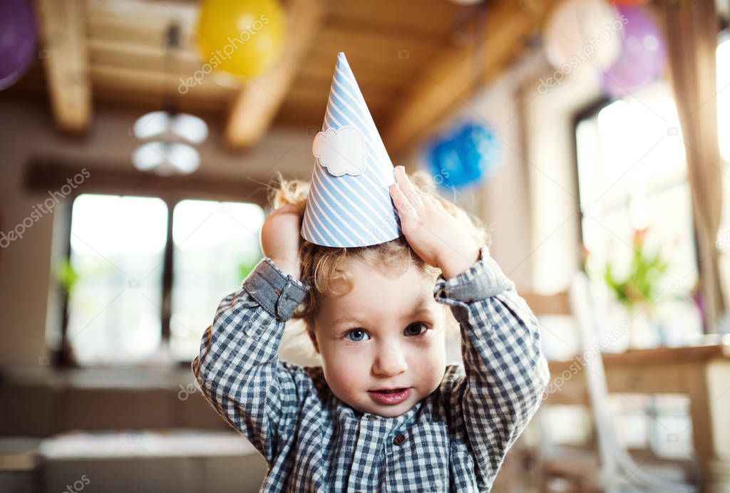 A toddler boy with a party hat standing inside at home.