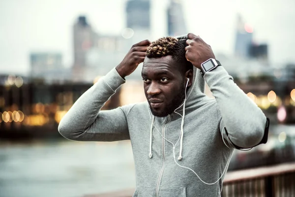 Fekete férfi futó fülhallgató a hídon, egy város, elhelyezés csuklya fej. — Stock Fotó
