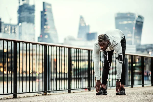 Black man runner with smartphone in an armband on the bridge in a city, stretching.