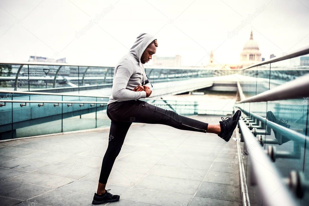 Black man runner with smartwatch on the bridge in a city, stretching.